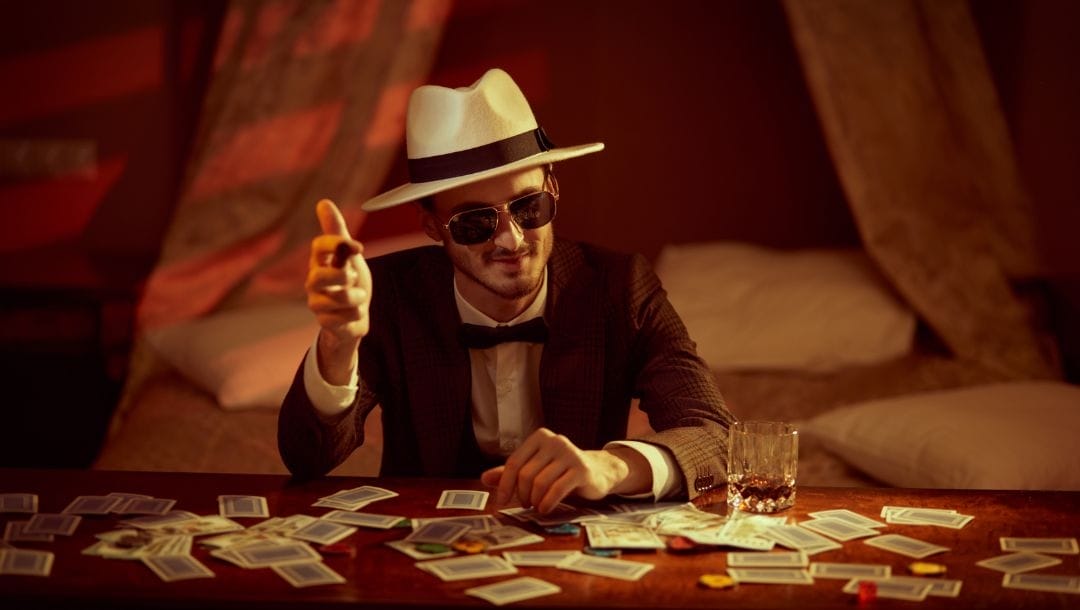 Header image, a man wearing a suit and hat sitting at a desk scattered with playing cards and poker chips as he points a cigar towards the camera, there is a bed in the background