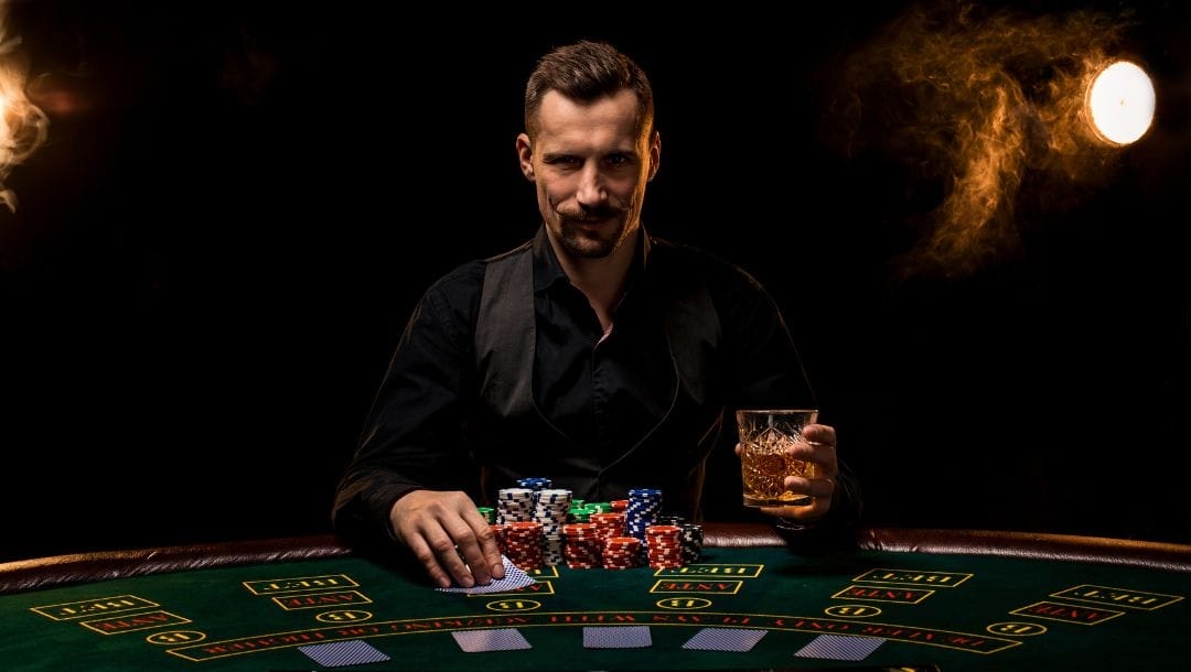 a man with a mustache sitting at a green felt poker table with poker chips stacked in front of him and a drink and playing cards in his hands