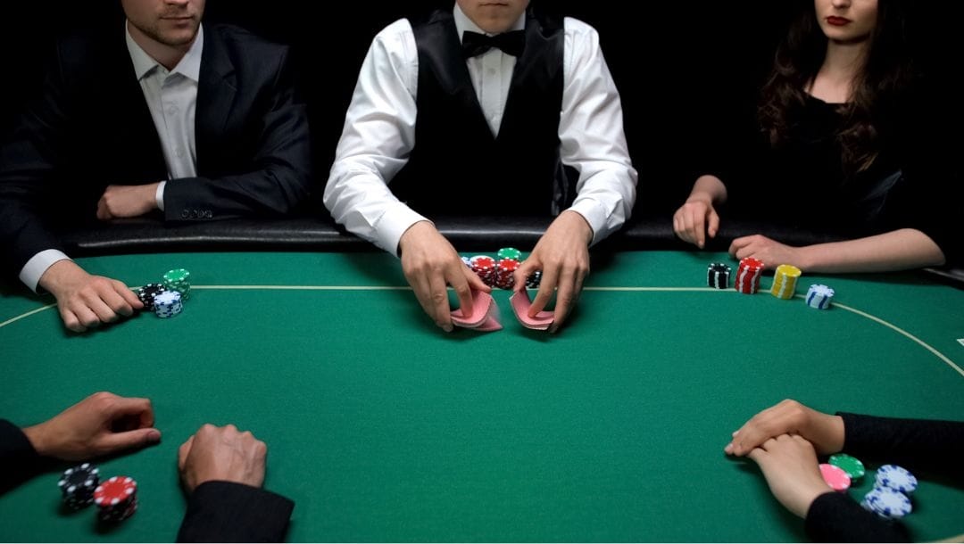 four poker players and a dealer, all in formal attire with poker chips stacked in front of them, sit around a green felt poker table as the dealer shuffles a deck of playing cards.