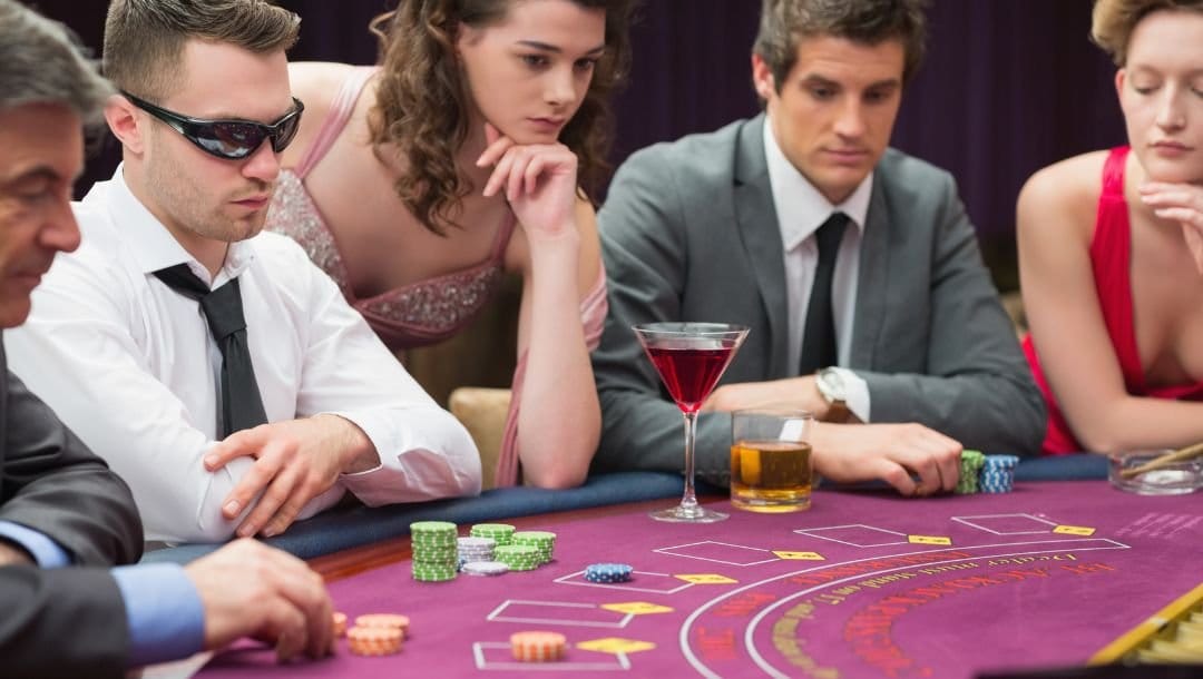 Five well dressed people sitting at a purple felt poker table playing poker waiting for cards to be dealt, two drinks sit on the table