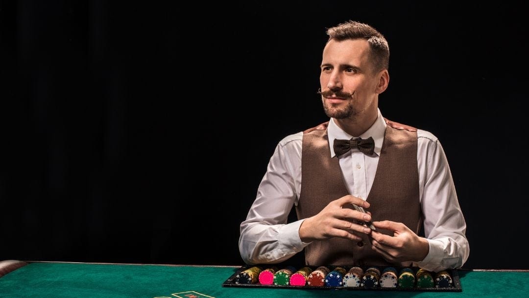 a man with a mustache in a vest and bowtie sitting at a green felt poker table behind poker chips looking into the distance