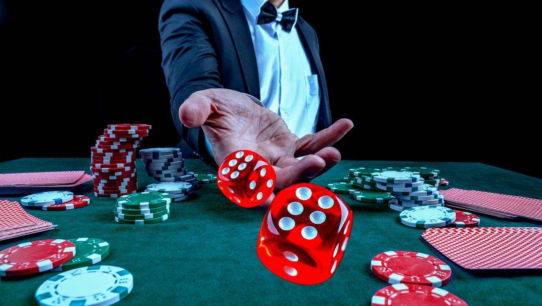 a man in a suit and bowtie throwing a pair of red dice forward on a green felt poker table stacked with poker chips and playing cards