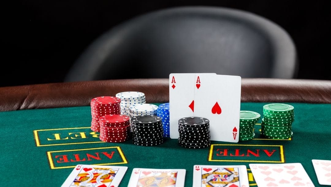 a green felt blackjack table set up in a casino with playing cards and stacks of poker chips, a pair of red aces stand against the poker chips and a black chair in the back