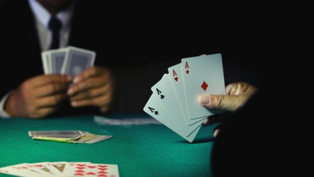 two players sitting opposite each other at a green felt poker table holding playing cards, the hand facing the camera being four of a kind aces