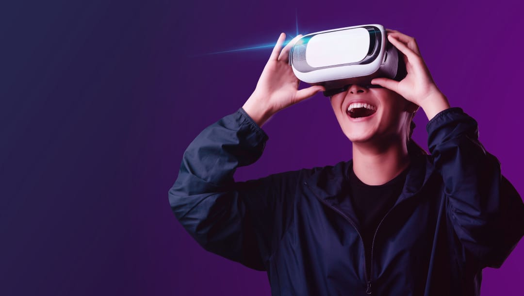A photograph of a woman standing against a purple background wearing VR glasses and smiling with her hands gently gripping the sides of the headset.