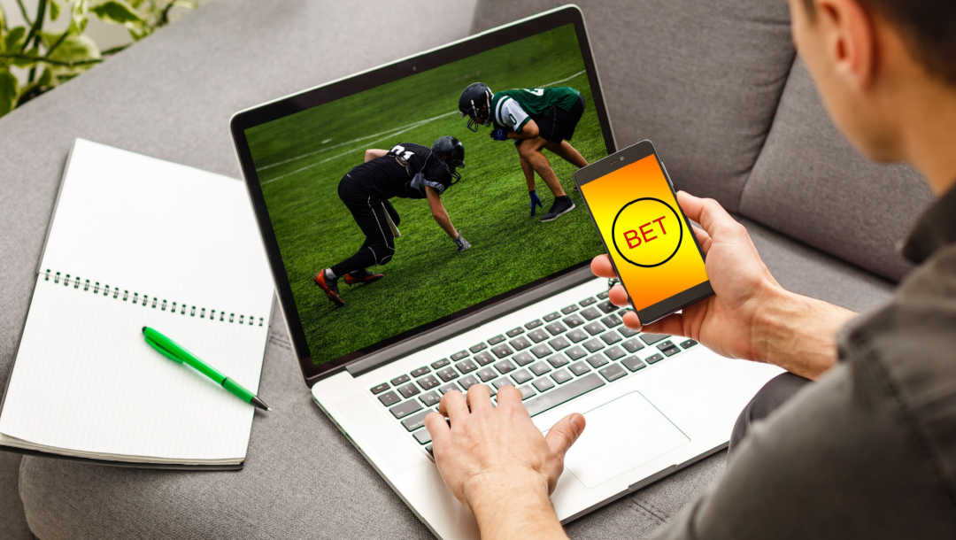 A person watching an American football game on their laptop places a bet on their mobile phone.