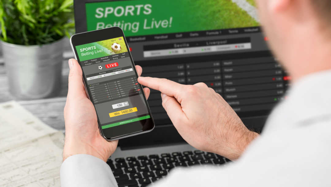 A person uses their phone and laptop with sports betting open on the screens.