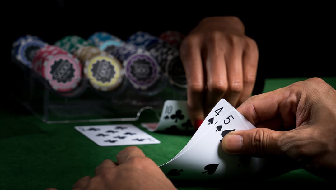 Close-up of a Chemin de Fer game in progress as a player peeks at their cards, revealing a four and a five of spades, with colorful chips in a chip rack in the background. The dealer is revealing their second card.