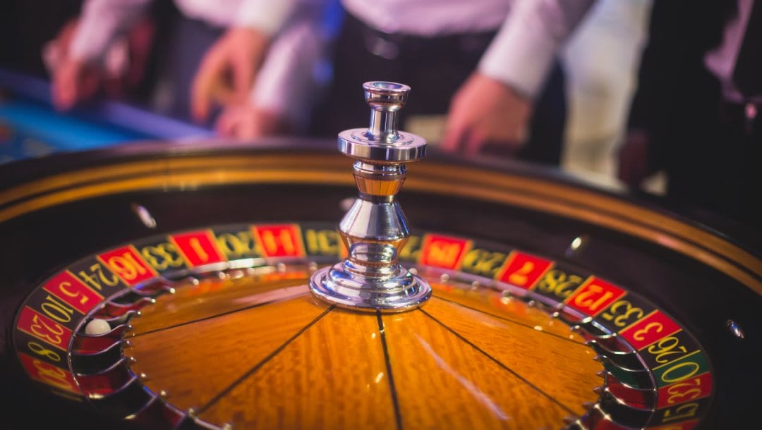 A closeup of a roulette wheel with gamblers in the background.