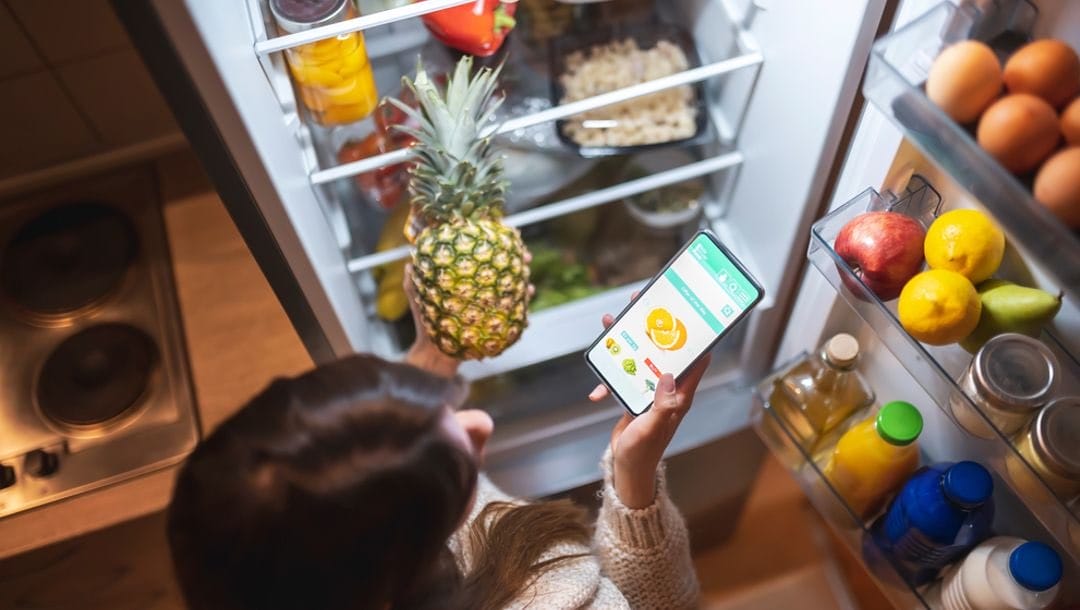 A woman standing at her fridge with her smartphone in one hand and a pineapple in the other.