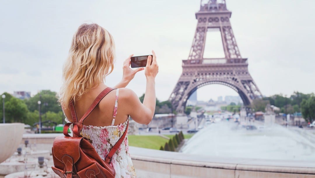 A blonde tourist in a floral dress with a leather backpack taking a photo of the Eiffel Tower on her smartphone.