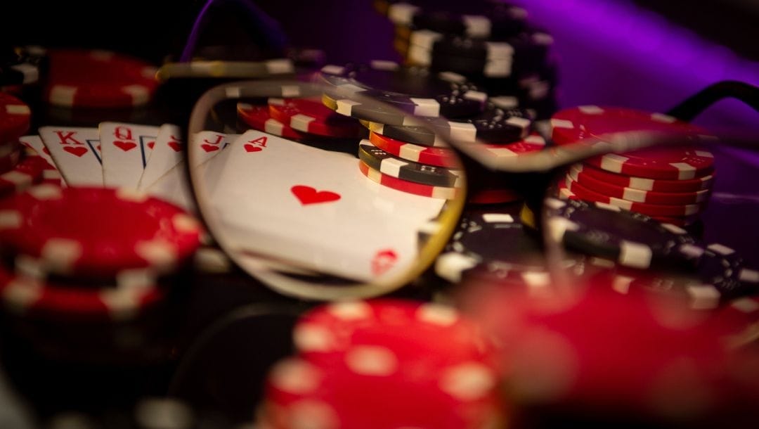 a pair of glasses magnifying a royal flush of hearts hand of playing cards on a table that has stacks of red and black poker chips on it