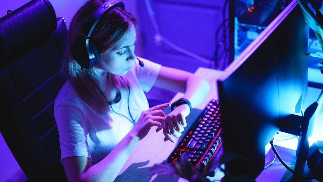 A female gamer sitting in front of her computer looks at her watch.