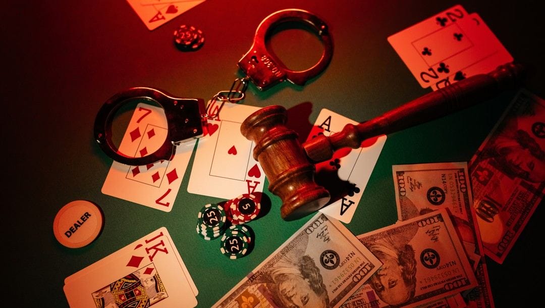 A gavel, handcuffs, playing cards, casino chips and cash scattered on a green casino table. The items on the table are bathed in red light.