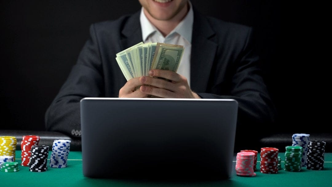 A man wearing a suit sits in front of his laptop with poker chips stacked on each side and cash in his hands.
