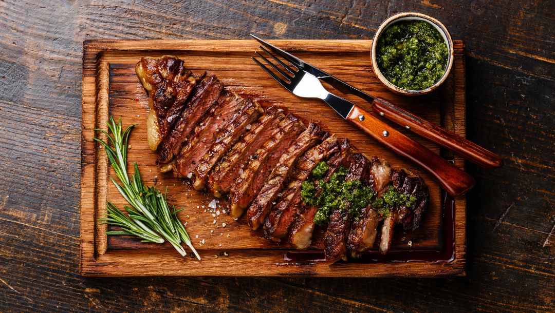 sliced grilled beef barbecue striploin steak with chimichurri sauce on a wooden cutting board on dark wooden background