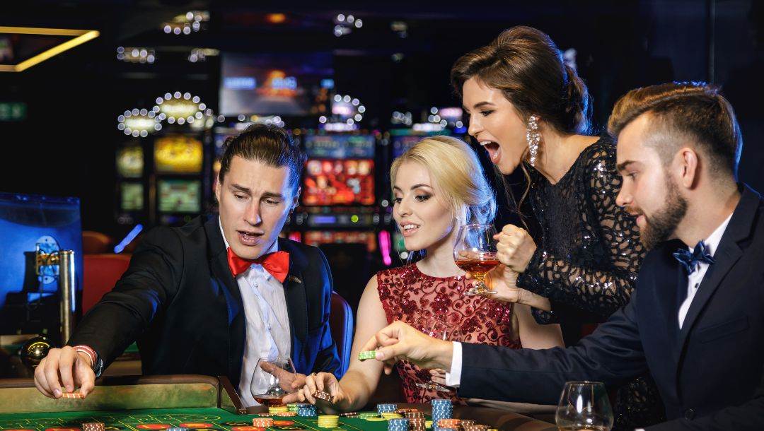 four well dressed people, two men and two women, sitting at a roulette table placing bets and cheering each other on in a casino