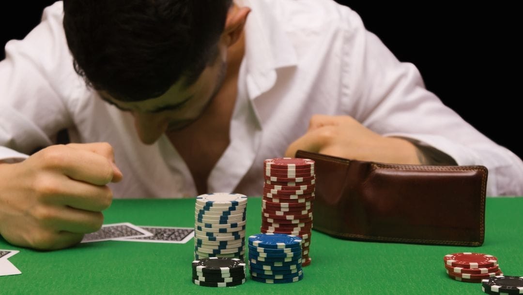 a man in distress sitting at a poker table with playing cards, poker chips and a wallet in front of him on the table