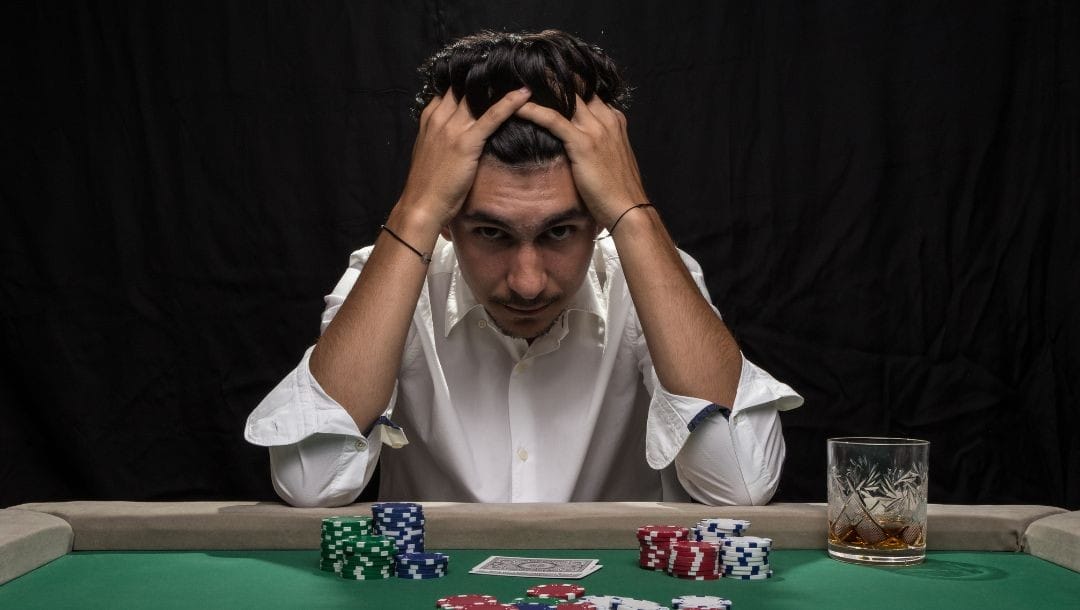 a man sitting at a poker table with his head in his hands in distress with playing cards, poker chips and a drink glass on the table in front of him