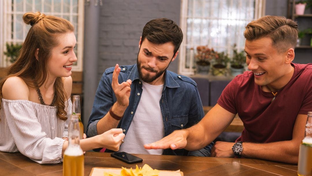 three friends, a woman and two men, are sitting at a dining table playing rock, paper, scissors - there are drinks, food and a cellphone on the table