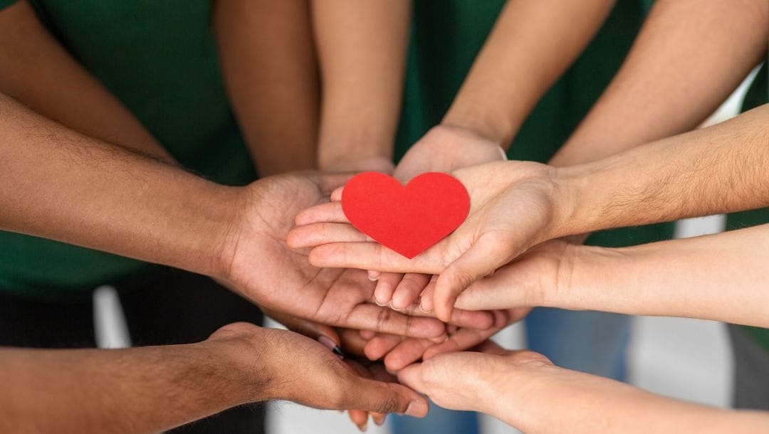 multiple people standing in a circle, a close up of their hands with palms facing up on top of eachother and a red cardboard heart on top