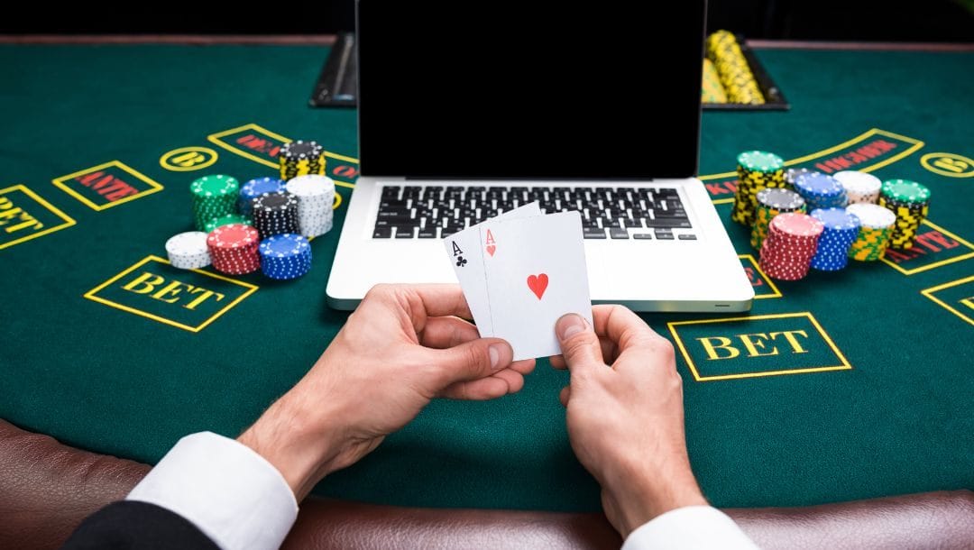 a man’s hands holding a pair of ace playing cards while sitting at a poker table, on the table is an open laptop and stacks of poker chips on either side of it