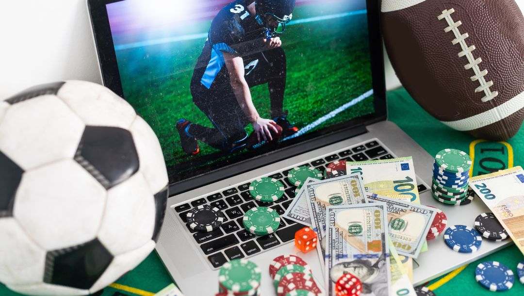 online sports betting and gambling concept of a laptop displaying a football player during a game, the laptop is on a green felt poker table with a soccer ball and football on either side of it, poker chips, red six-sided dice and money notes are scattered on the laptop keyboard