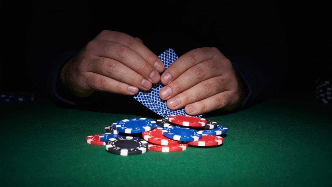 a close up of a man’s hands holding two playing cards coming out of the shadow towards the poker chips on the green felt poker surface