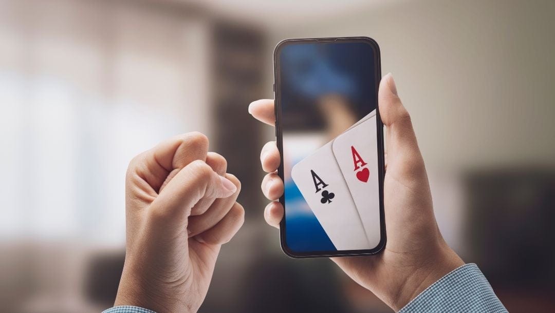 a person holding up a cellphone with online poker on it displaying a pair of ace playing cards, the other hand is in a fist