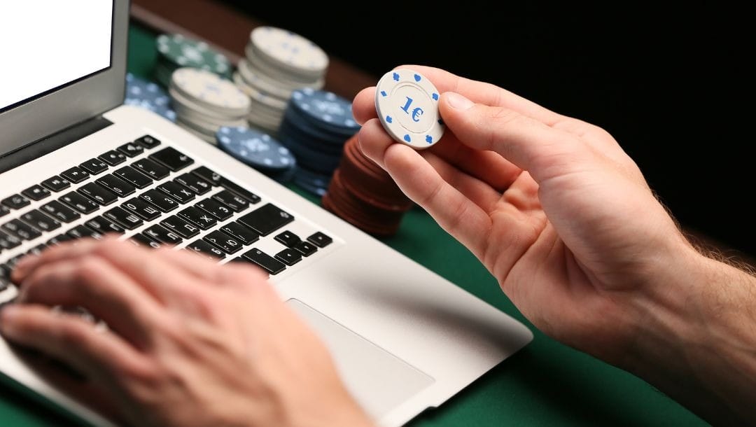 a person typing on a laptop with their left hand and holding a poker chip with their right hand above a green felt poker surface stacked with poker chips
