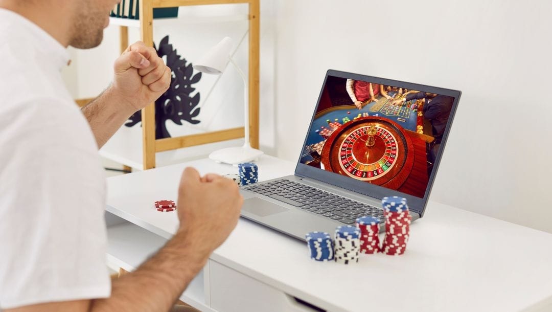 a man playing online roulette on a laptop that’s on a white desk with poker chips stacked on either side of the laptop