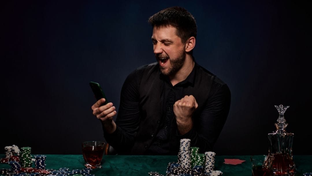 a man cheering while holding a cellphone and sitting at a green felt poker table that has stacks of poker chips, playing cards and drinks on it