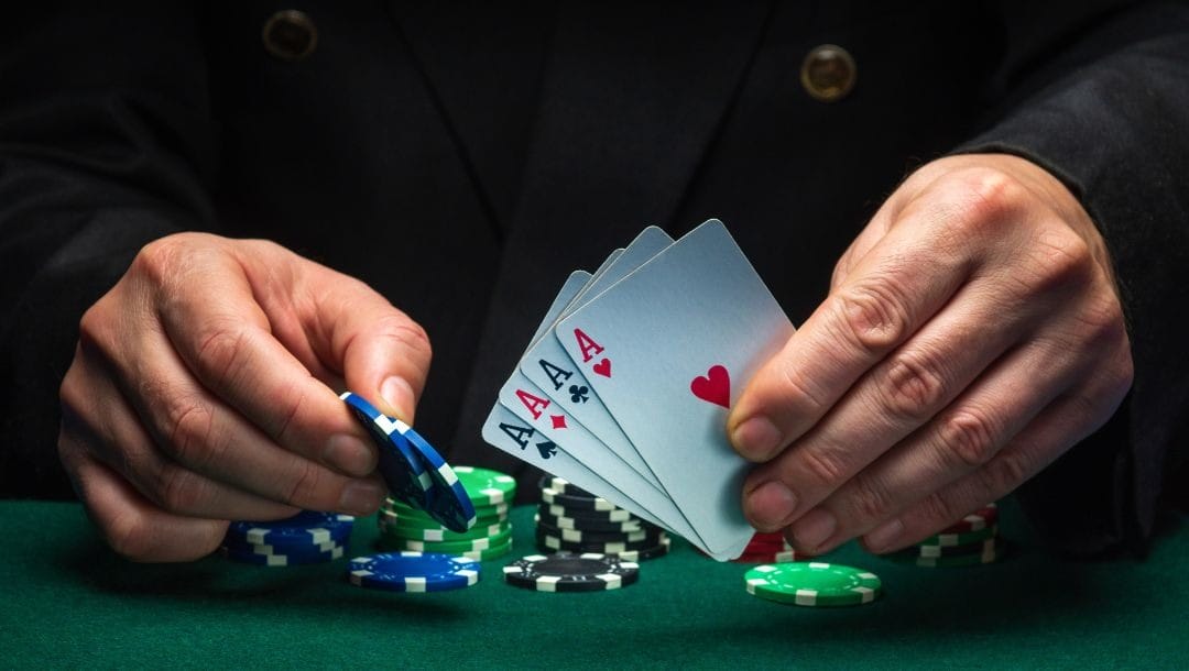 a man holds four of a kind ace playing cards in his left hand and two blue poker chips in his right hand over a green felt poker table with poker chips stacked on it