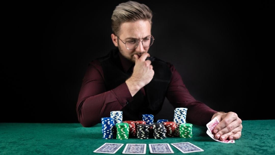 a man is sitting at a green felt poker table with stacks of poker chips and four playing cards laid out face down in front of him, he is looking inquisitively at his hole cards in his left hand