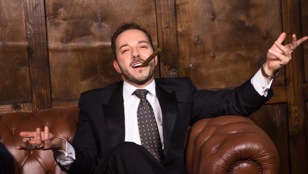 a man wearing a suit with a cigar in his mouth is sitting and shrugging on a leather sofa with wooden panel walls behind him
