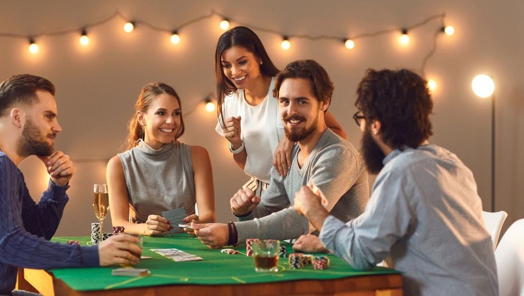 five friends are playing poker while gathered around a dining table that has been turned into a poker table with a green felt table cover, playing cards and poker chips on it, in the background are hanging lights on the wall