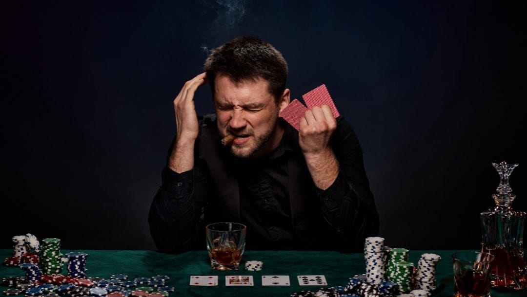 a man in distress is smoking a cigar while playing poker and holding two hole cards, on the table are stacks of poker chips, playing cards and a whiskey glass