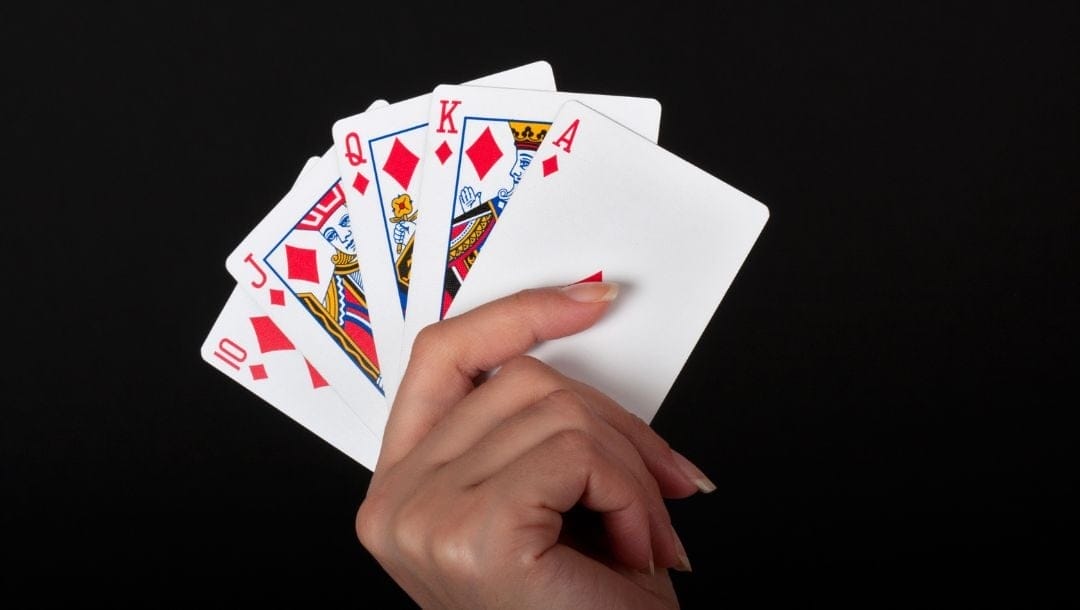 a woman’s hand holding up five playing cards, a royal flush of diamonds, with a pitch black background
