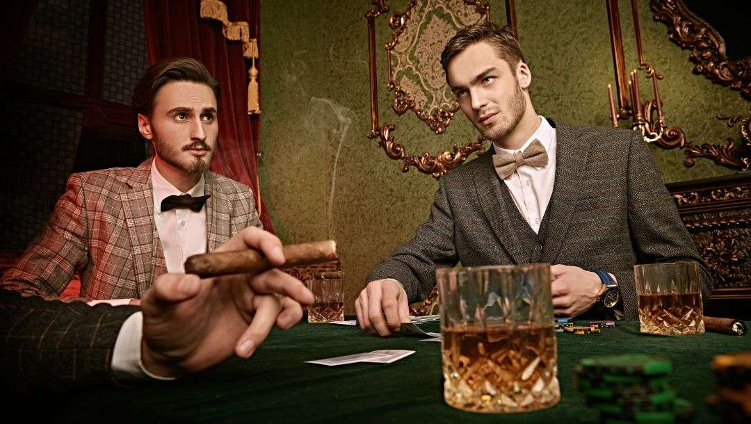 two men in suits and another with only his hand holding a cigar in the frame, all sitting around a poker table in a vintage style room playing poker and drinking whiskey out of crystal whiskey glasses
