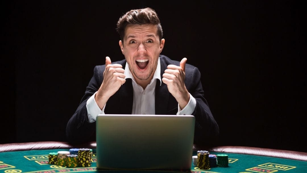 a very happy man with his thumbs up is sitting at a poker table with a laptop open and poker chips stacked in front of him