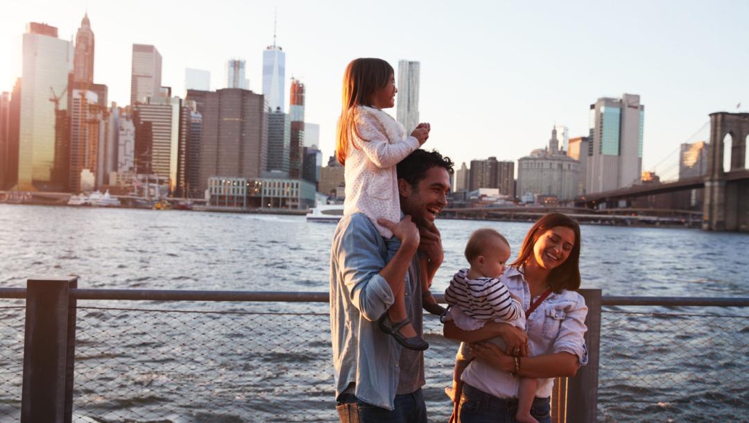 A young family standing by the river’s edge with a cityscape in the background.