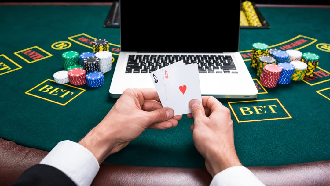 A close-up of a man’s hands resting on a blackjack table, holding two ace cards. There is a laptop in front of him with stacks of casino chips on each side of it.