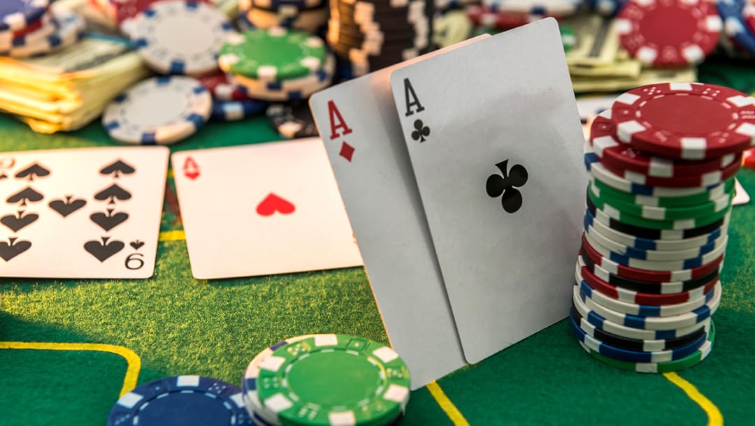 A pair of aces leaning against stacks of casino chips. An ace and a nine are on the table behind the pair of aces. Stacks of casino chips and banknotes are scattered around the green casino table.