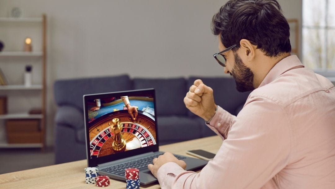 A man holding his fist in a gesture of victory while looking at a laptop displaying online roulette.