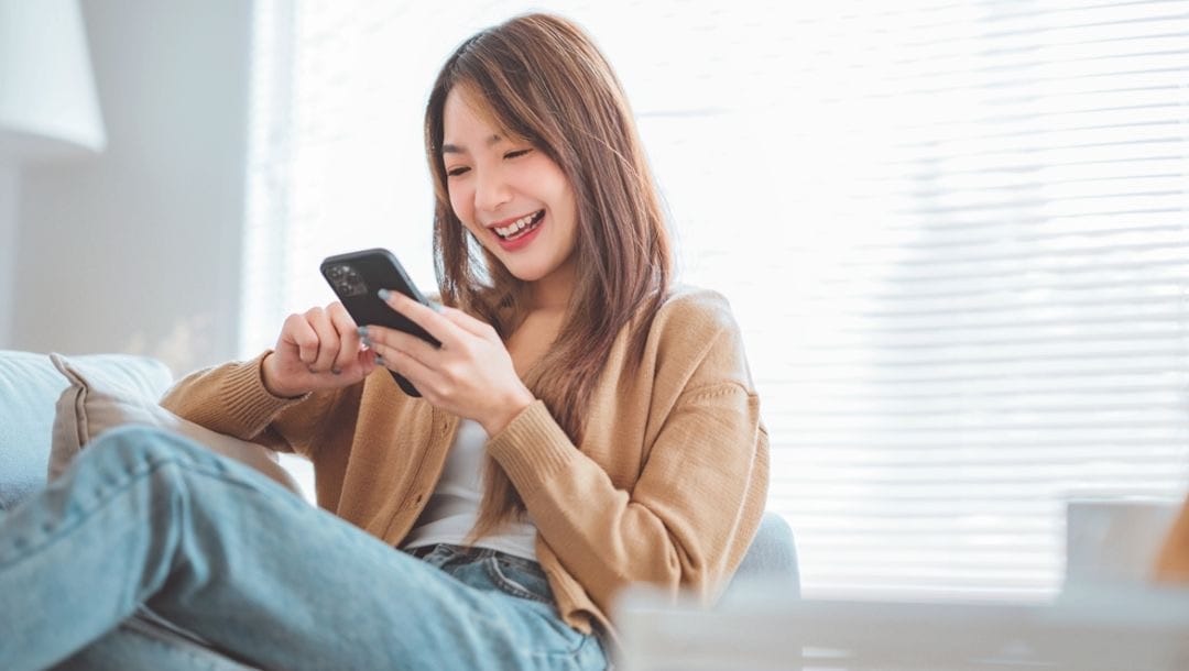 A woman smiling as she looks at her smartphone while sitting on a couch.