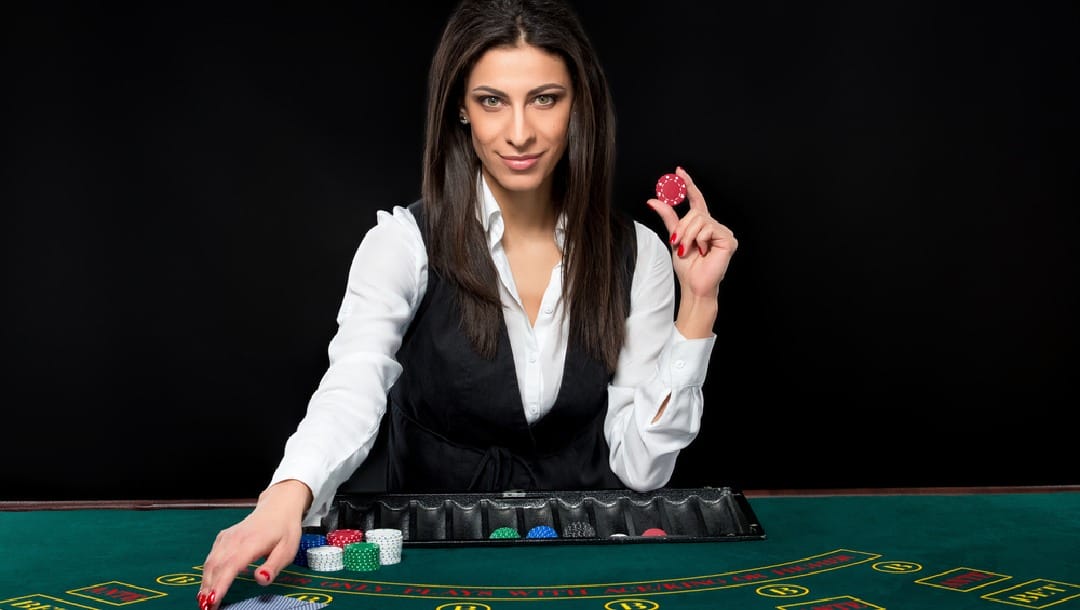 A woman poker dealer holding a poker chip by a green felt table.