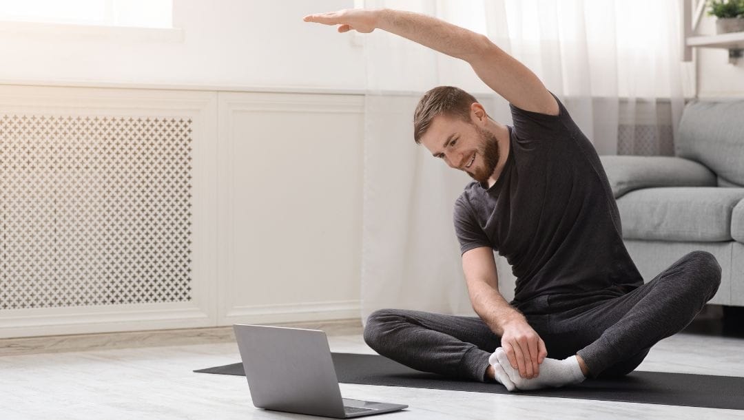 a man on a yoga mat on the floor is stretching to a yoga tutorial on a laptop in front of him