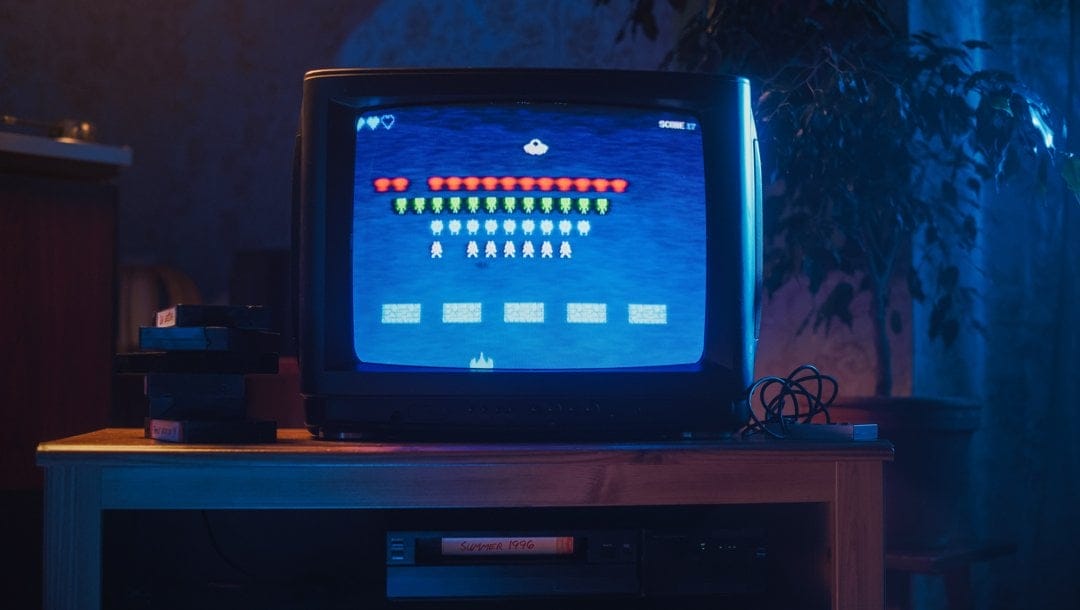 A CRT TV on a table with a game that looks similar to Space Invaders on screen. A controller sits next to the TV with VHS tapes on the other side.