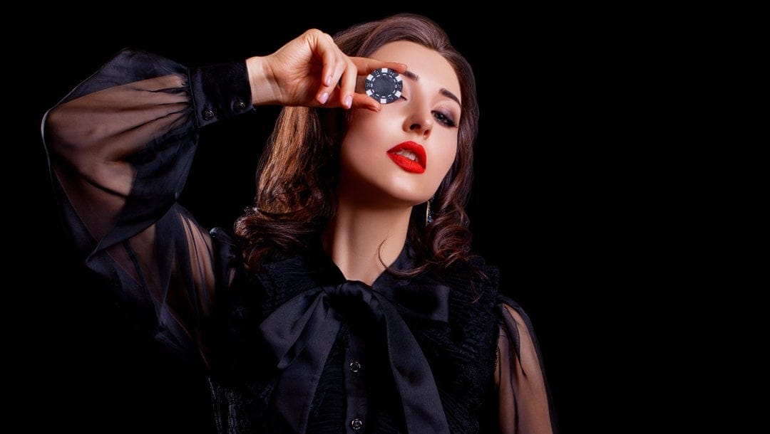 A woman in a black dress holds a casino chip up to her eye.