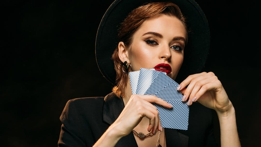 A beautiful woman wearing a fedora and a suit jacket holds a hand of five playing cards.